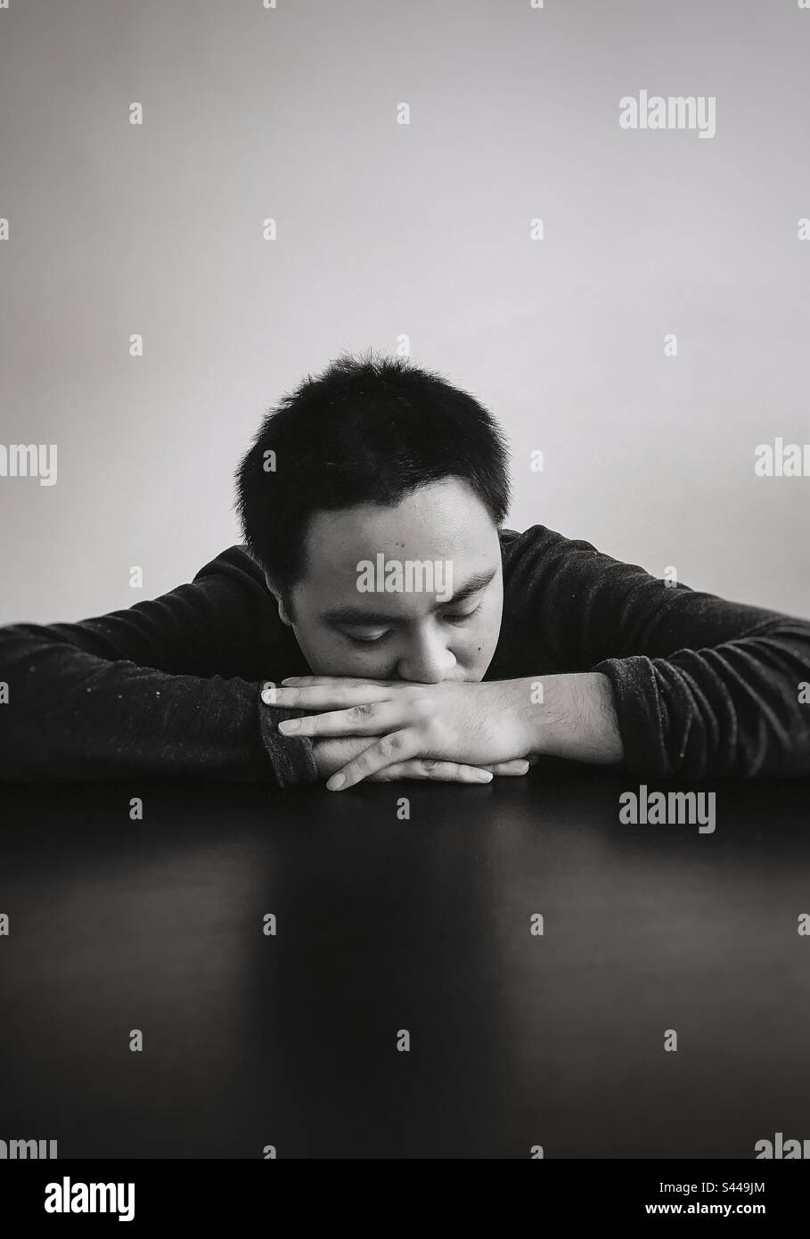 Monochrome portrait of young Asian man with eyes closed and head resting on folded arms on table. Copy space. Emotion. Sadness. Tiredness. - Smartphone Captured Stock Image