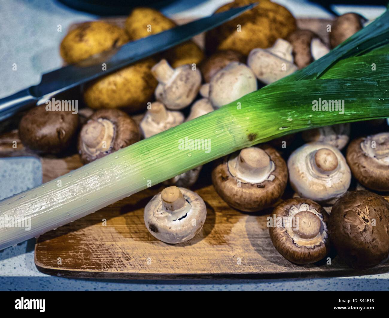 The makings of cream of mushroom, leek and potato soup. Brown mushrooms, white button mushrooms, leek and Dutch cream potatoes on wooden cutting board with kitchen knife on countertop. - Smartphone Captured Stock Image