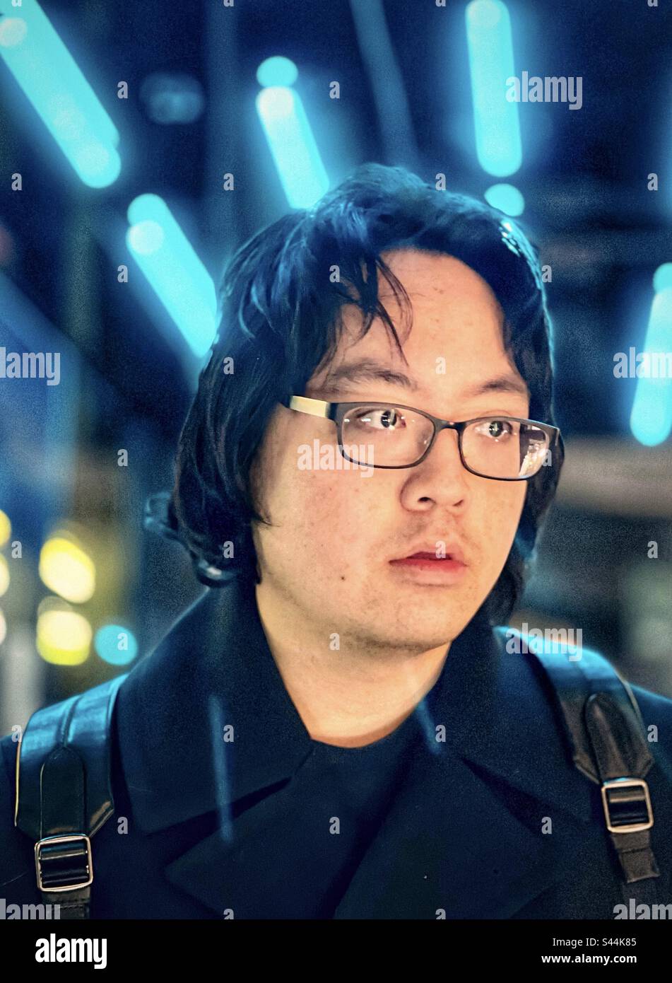 Close-up portrait of young Asian man in eyeglasses standing against blue neon lights in the city at night. - Smartphone Captured Stock Image