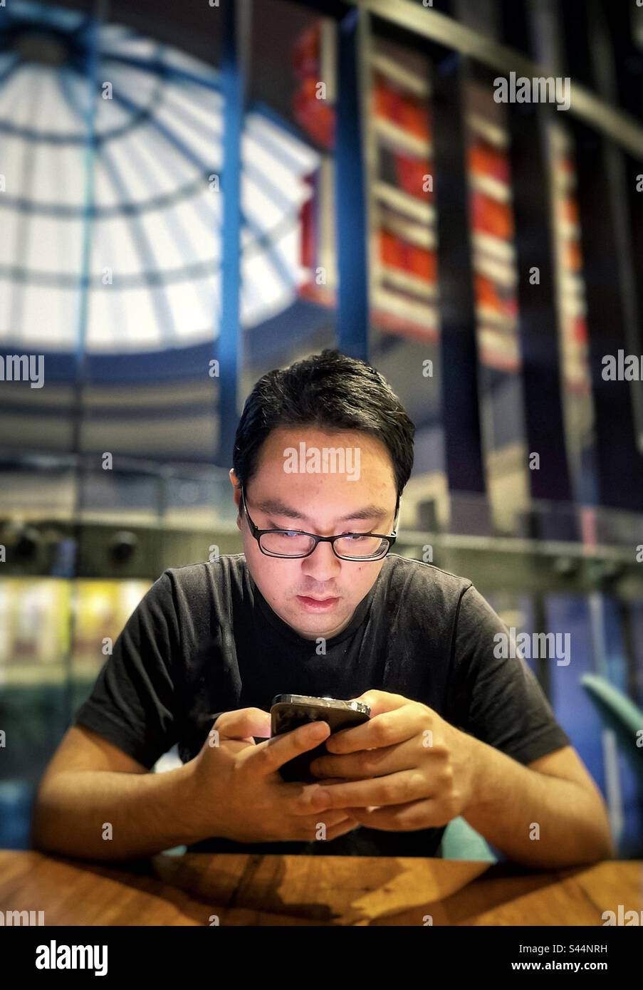 Young Asian man in eyeglasses using mobile phone at table inside cafe against architectural features and skylight. - Smartphone Captured Stock Image