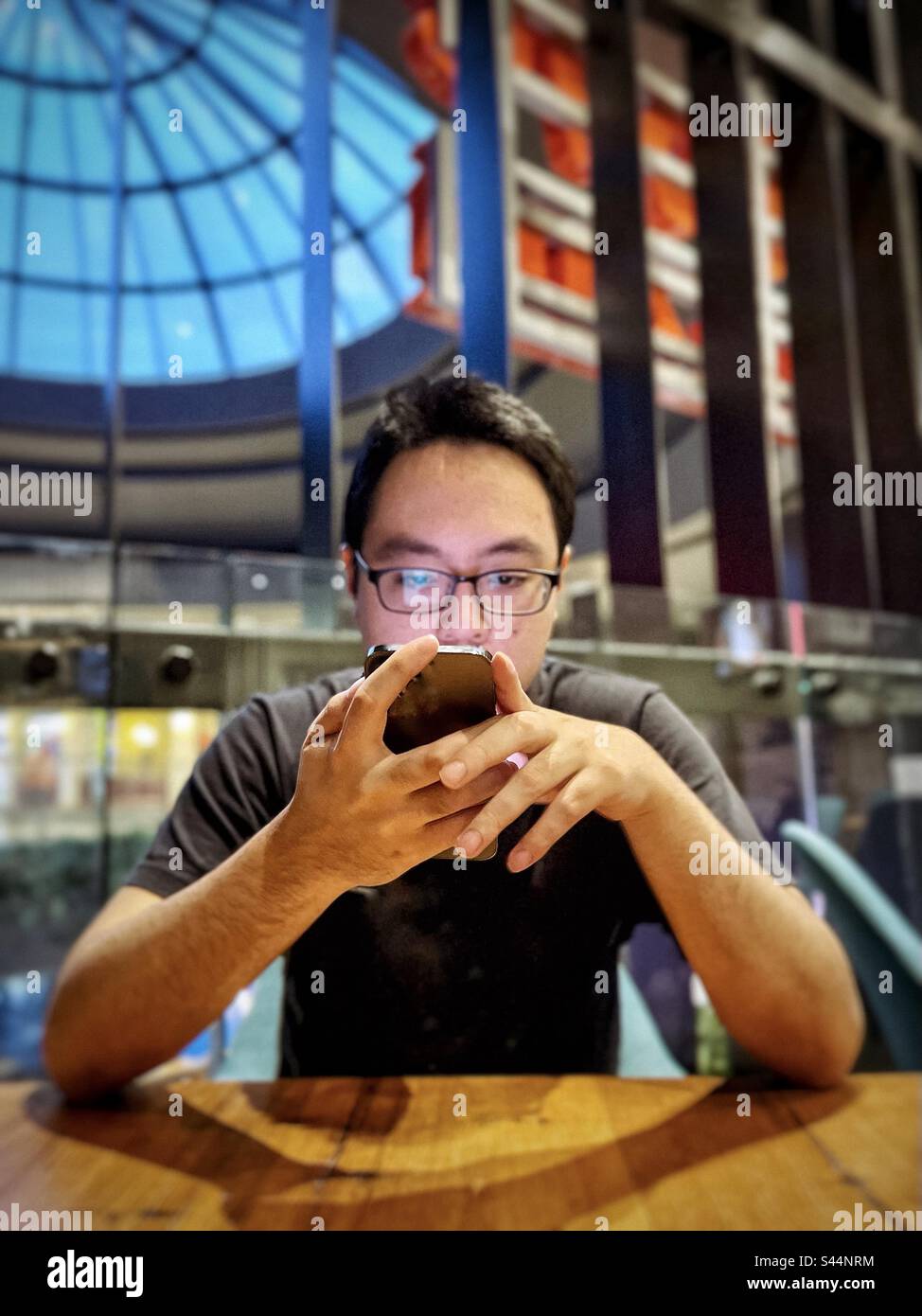 Portrait of young Asian man using mobile phone at table inside cafe against architectural features and skylight. - Smartphone Captured Stock Image