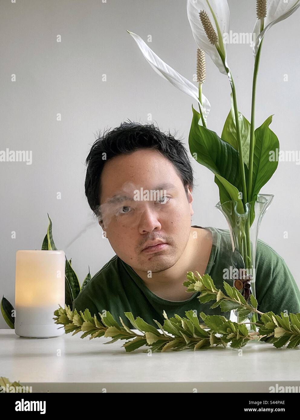 Young Asian man inhaling essential oils from aromatherapy diffuser, surrounded by peace lilies in vintage vase and green branches on table. Aromatherapy. Alternative therapy. Well-being, self care. - Smartphone Captured Stock Image