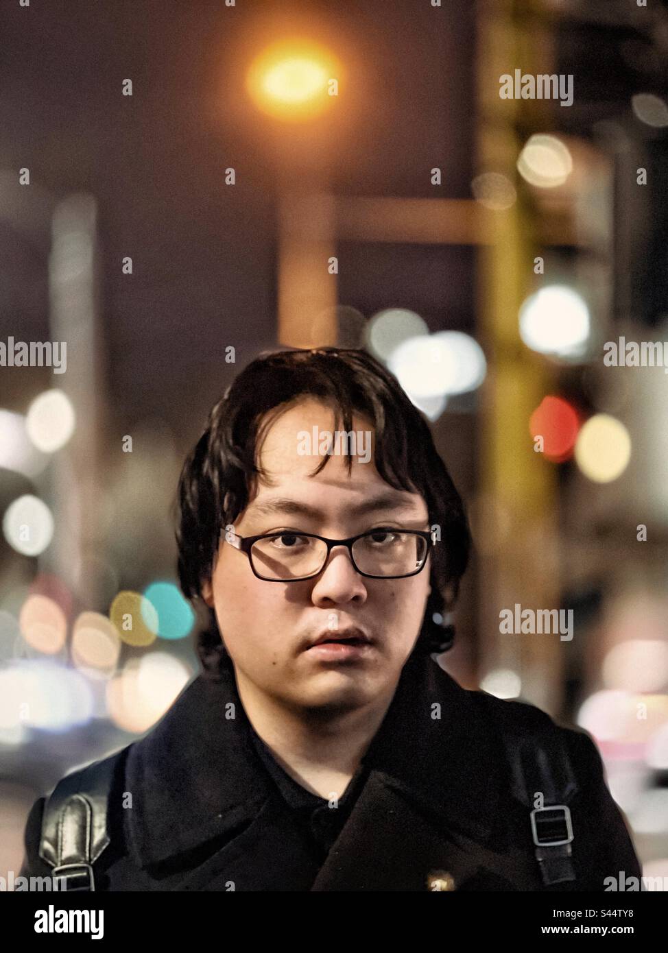 Portrait of young Asian man in eyeglasses against street lights and building in the city at night. Focus on foreground. - Smartphone Captured Stock Image