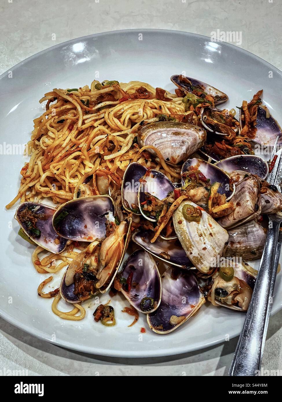 Close-up of spicy stir-fried noodles with pipis/clams on white plate with cutlery. Fusion cuisine. - Smartphone Captured Stock Image