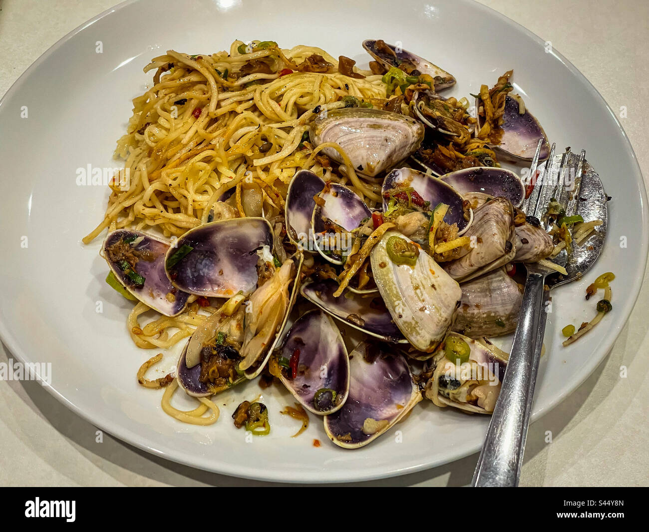 Close-up of spicy, stir-fried noodles with pipis/clams on white plate with cutlery on table. Fusion cuisine. - Smartphone Captured Stock Image