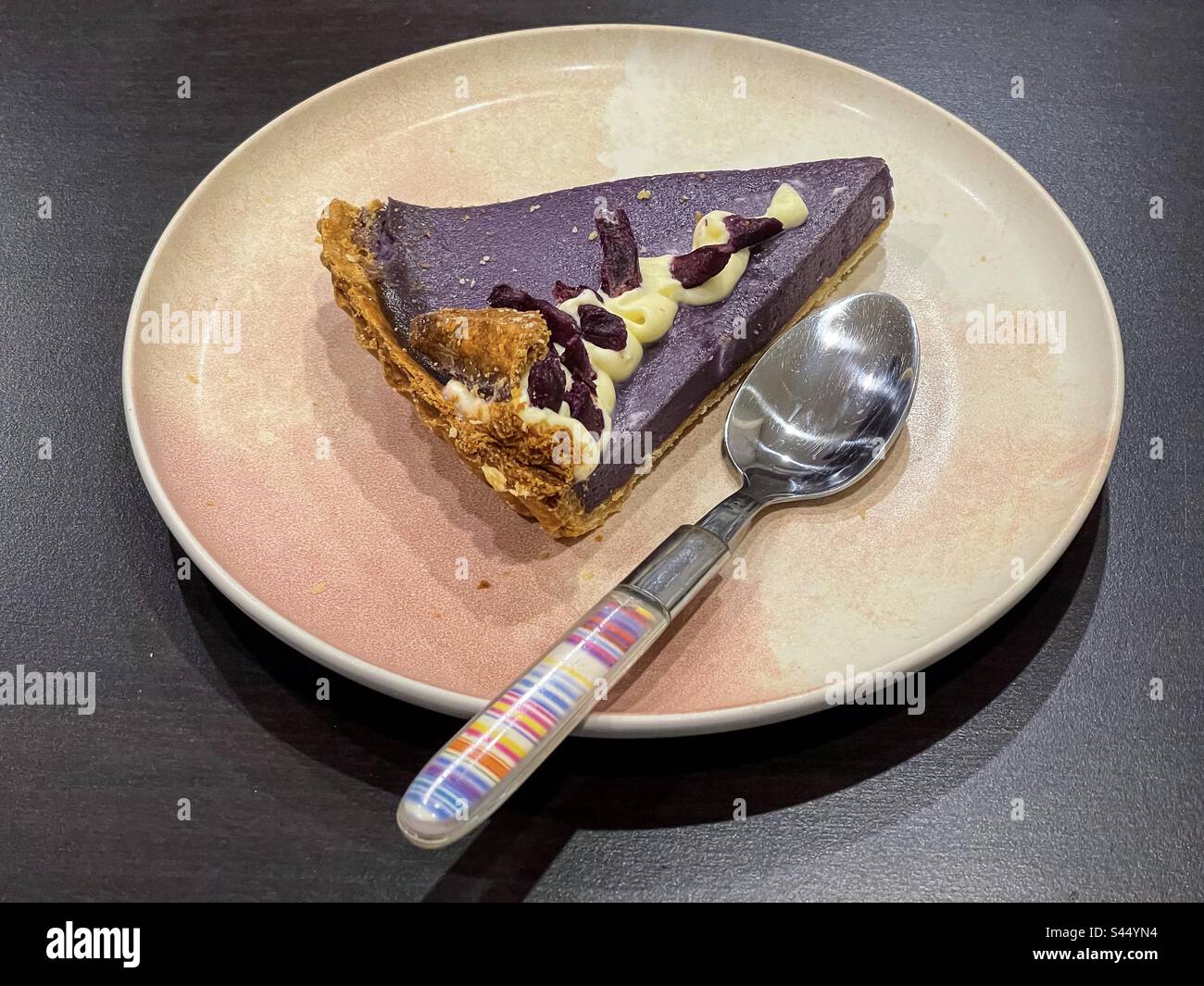High angle view of a slice of ube/purple yam pie with custard topping and purple yam pieces on plate with a spoon on table. Traditional Filipino dessert. - Smartphone Captured Stock Image