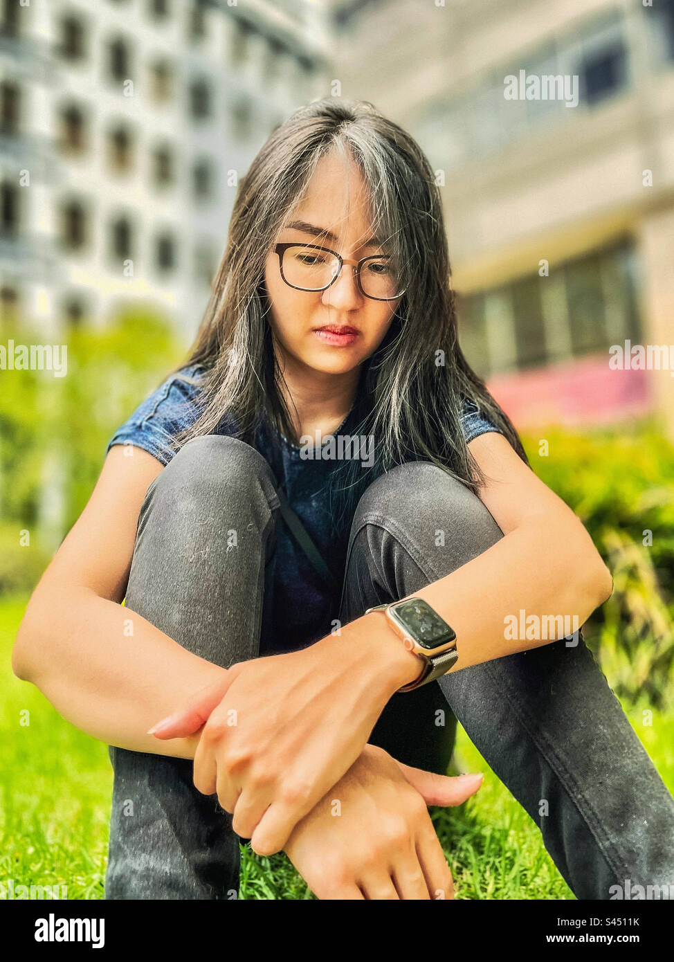 Emo girl. Portrait of young, long haired woman sitting on the grass with arms around her knees. Focus on foreground. Stock Photo