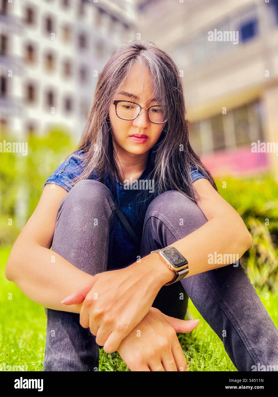Emo girl. Portrait of young, long haired, Asian woman in eyeglasses sitting on the grass with arms around her legs. Focus on foreground. Silver hair streaks. Stock Photo