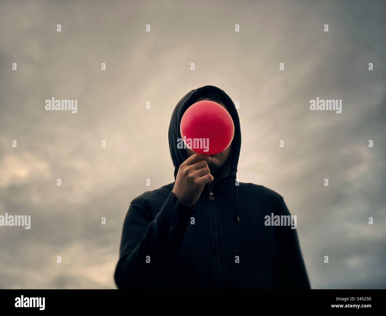Surreal man in black hoodie holding red balloon obscuring his face, standing against glowing cloudy sky. Dreamscape. Dystopian. - Smartphone Captured Stock Image