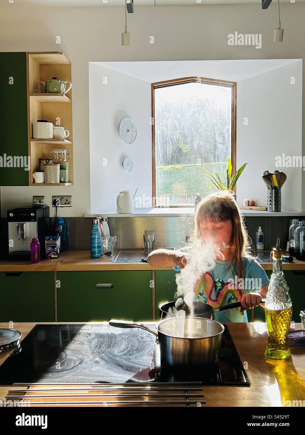 Girl cooking on a convection hob, stirring a pan with steam coming out. Window behind in an open plan kitchen Stock Photo