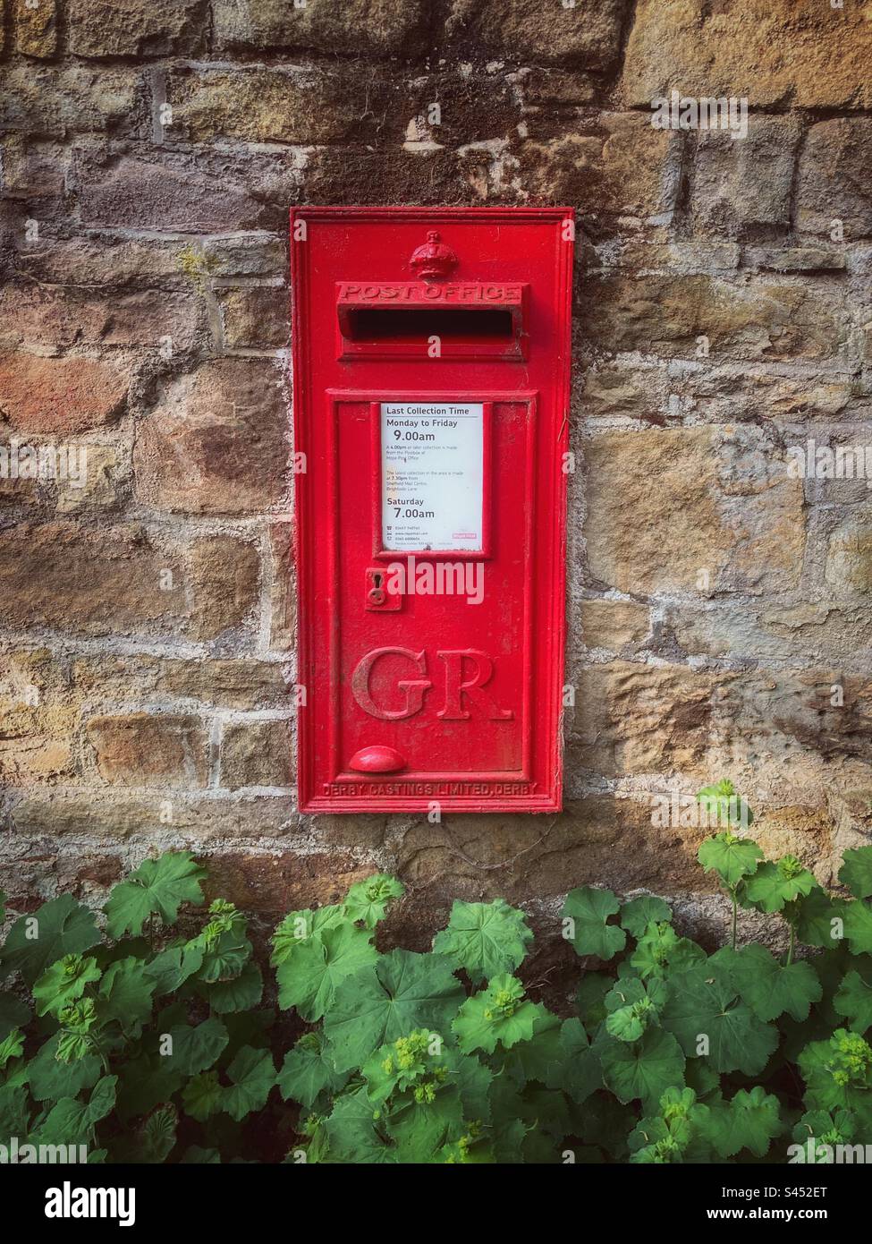 An old red post box set in a stone wall showing the letters GR for King George V. The white information sheet indicates this is an active post box with a daily Royal Mail collection. - Smartphone Captured Stock Image