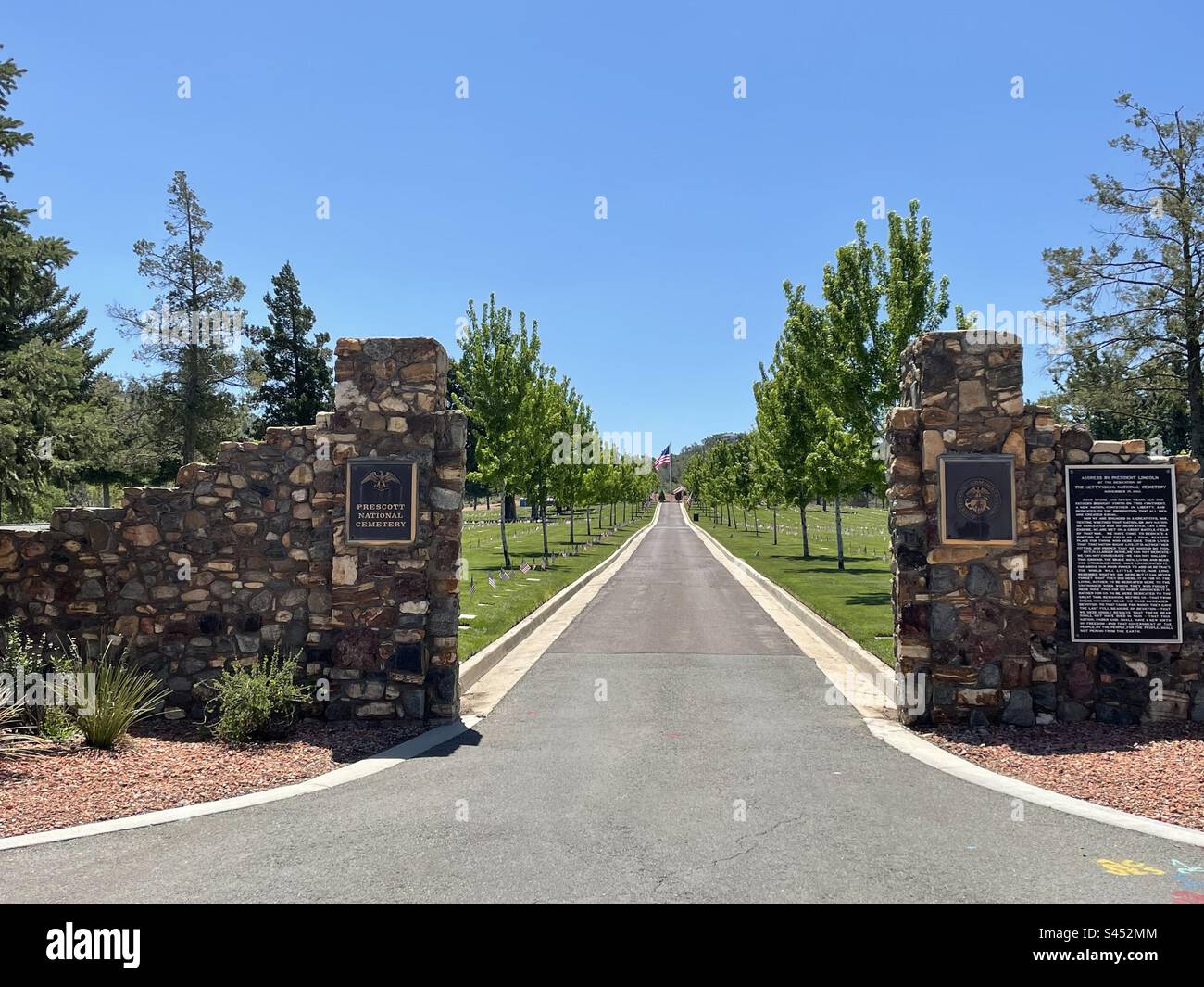 Prescott National Cemetery, historic entrance gate view, center drive leads up to American Flag, Veterans’ graves marked with American Flags, brilliant blue sky, framed by green trees, Arizona, USA Stock Photo