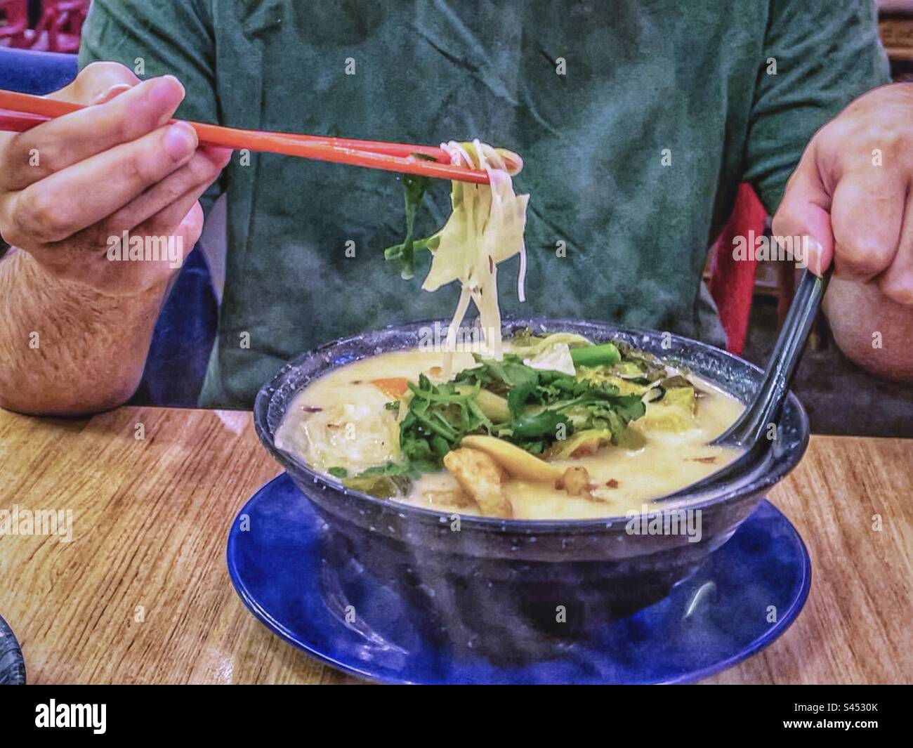 Midsection of man using chopsticks to lift vermicelli noodles from bowl of steaming hot vermicelli soup on table. - Smartphone Captured Stock Image