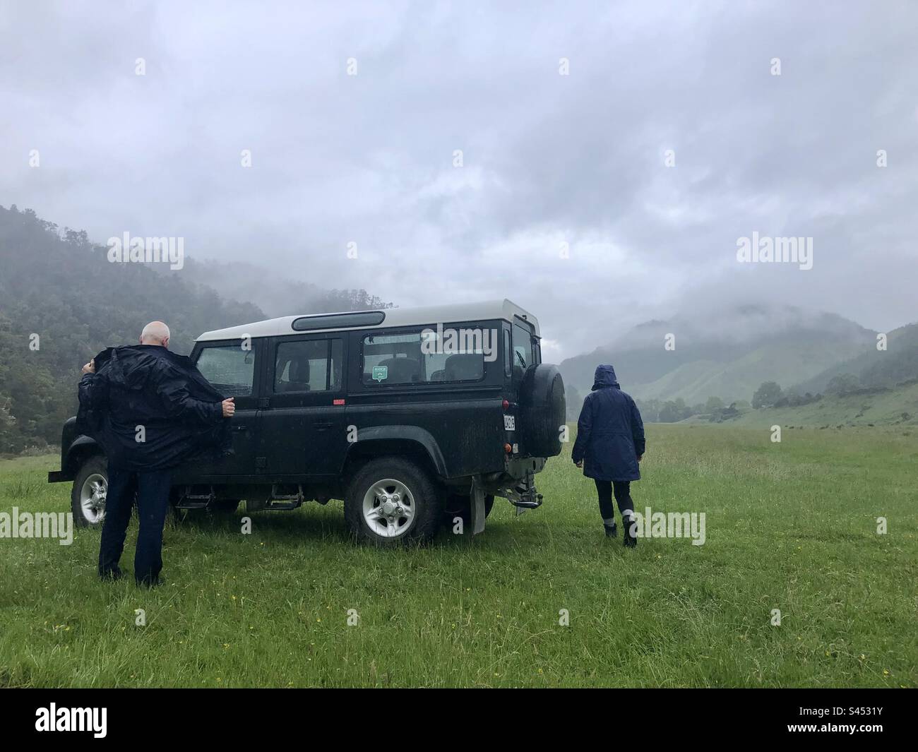 Misty day in the New Zealand hillside, two people in rain jackets are about to get into a Jeep. Stock Photo