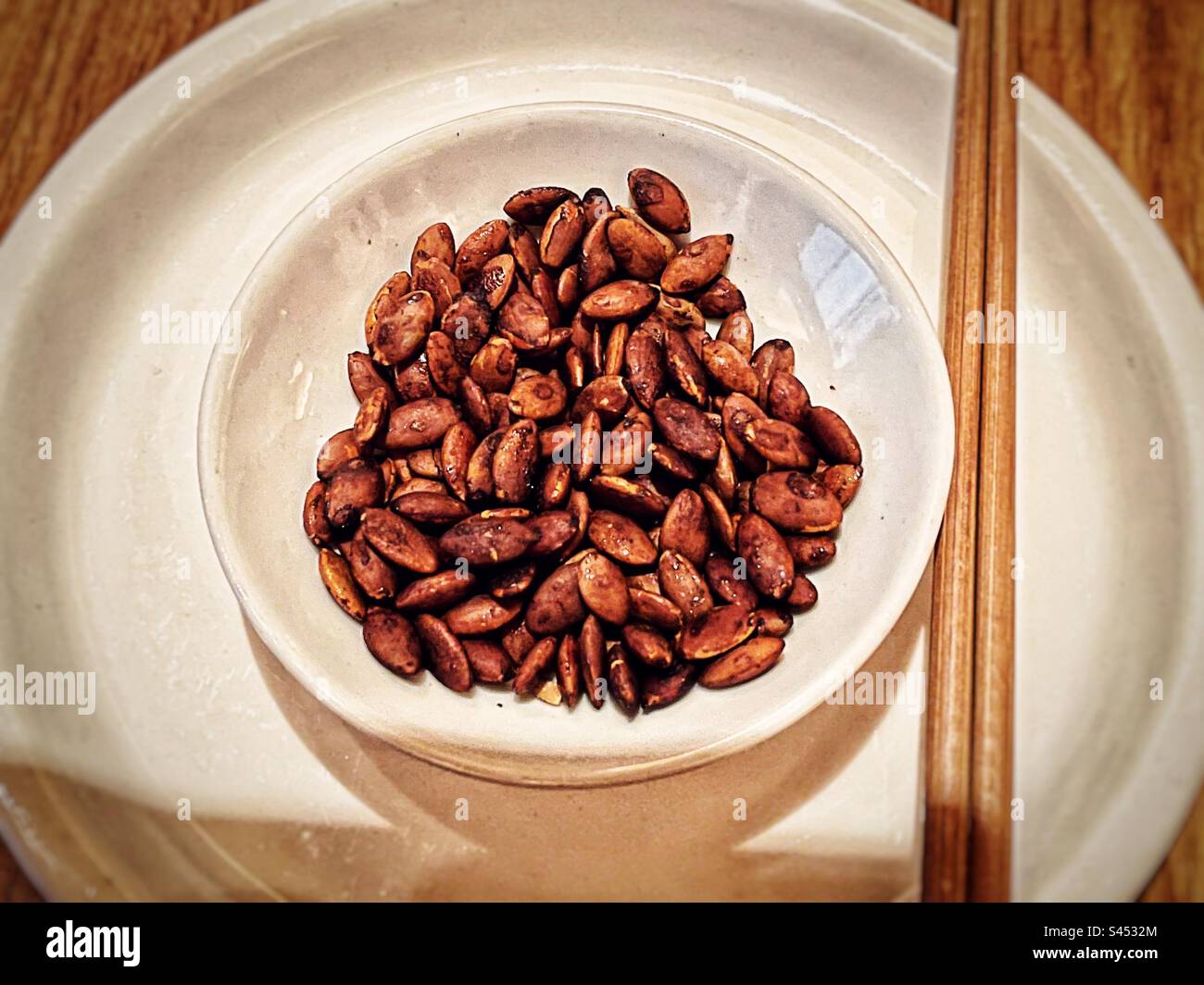 High angle view of seasoned pumpkin seeds in bowl on plate with chopsticks on table. - Smartphone Captured Stock Image