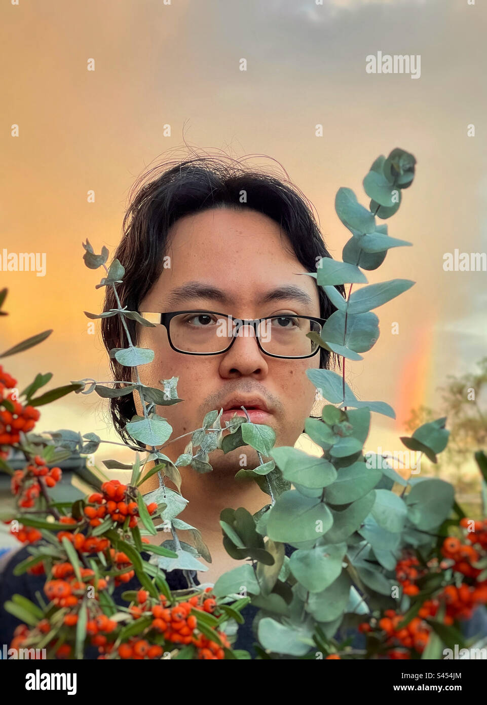 Portrait of young Asian man in eyeglasses with eucalyptus branches and orange Cotoneaster berries against sunset sky with rainbow. Stock Photo