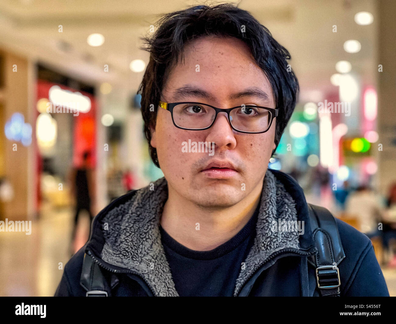 Close-up portrait of young Asian man in eyeglasses against lights inside shopping mall. Focus on foreground. Stock Photo