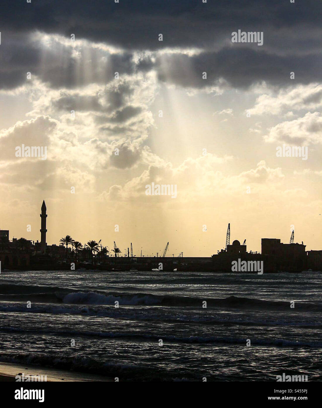 Crepuscular rays pass through a thick blanket of clouds to silhouette the port of Sidon in Lebanon and adjacent mosque. In the foreground are soft rolling waves. Stock Photo