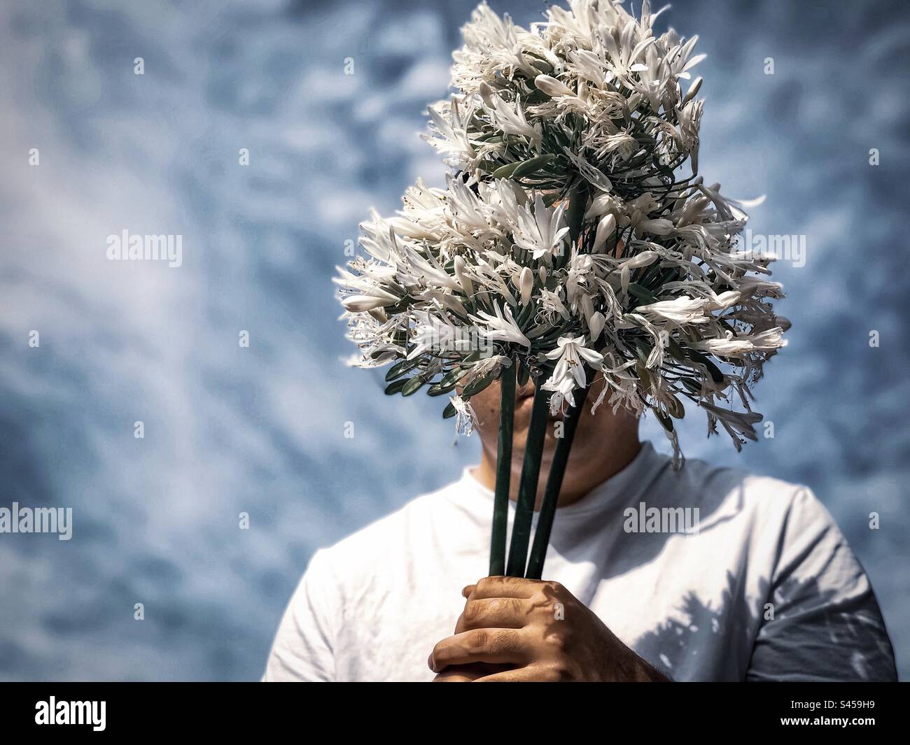 Portrait of young man in white T-shirt holding a bouquet of white Agapanthus africanus flowers aka African lily, Lily of the Nile against cloudy blue sky. Obscured face. Fashion. Spring theme. Stock Photo