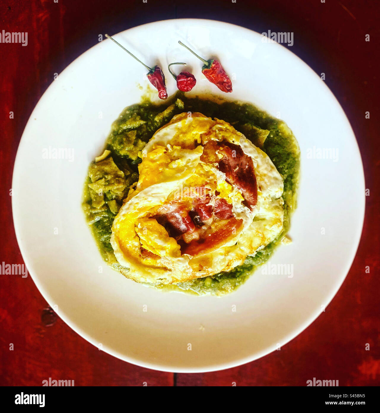 Fried eggs with bacon and three red hot chilli peppers on top of green chilaquiles in Queretaro, Mexico - Smartphone Captured Stock Image