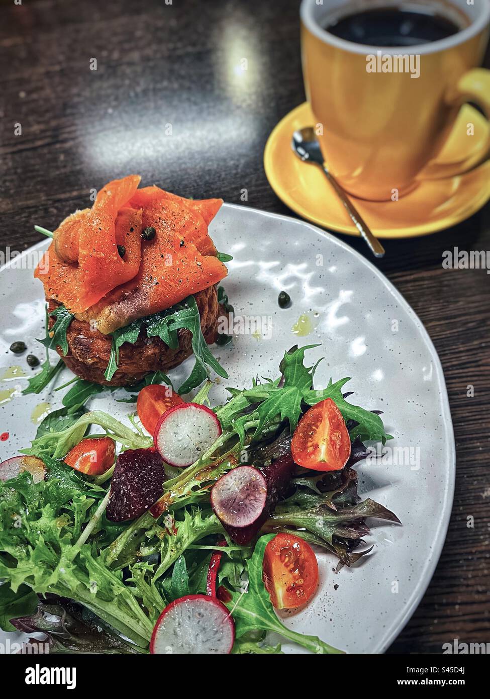 Smoked salmon on hot potato cakes with fresh salad on plate and a yellow cup of coffee on table. Stock Photo