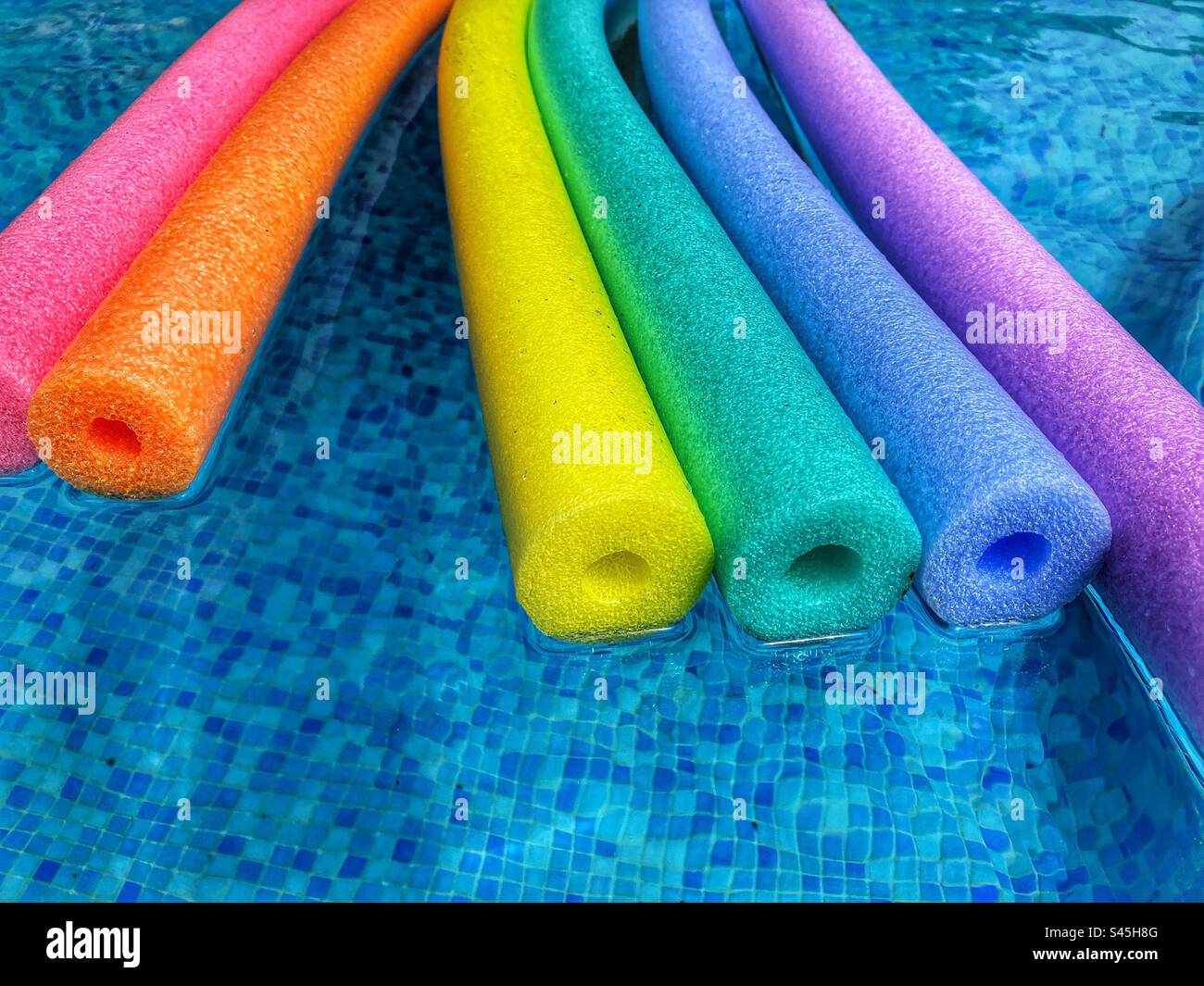 Summer vibes, rainbow coloured polyethylene pool noodles floating on water of blue tiled swimming pool, with copy space Stock Photo