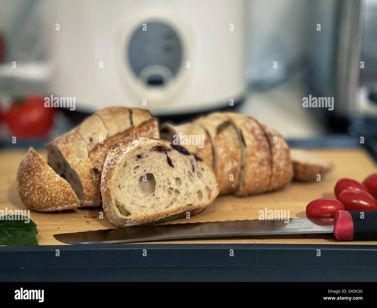 Close-up of sliced, crusty olive sourdough loaf of bread with cherry tomatoes, basil leaf and cutting knife on wooden cutting board  on kitchen countertop. Focus on foreground. - Smartphone Captured Stock Image