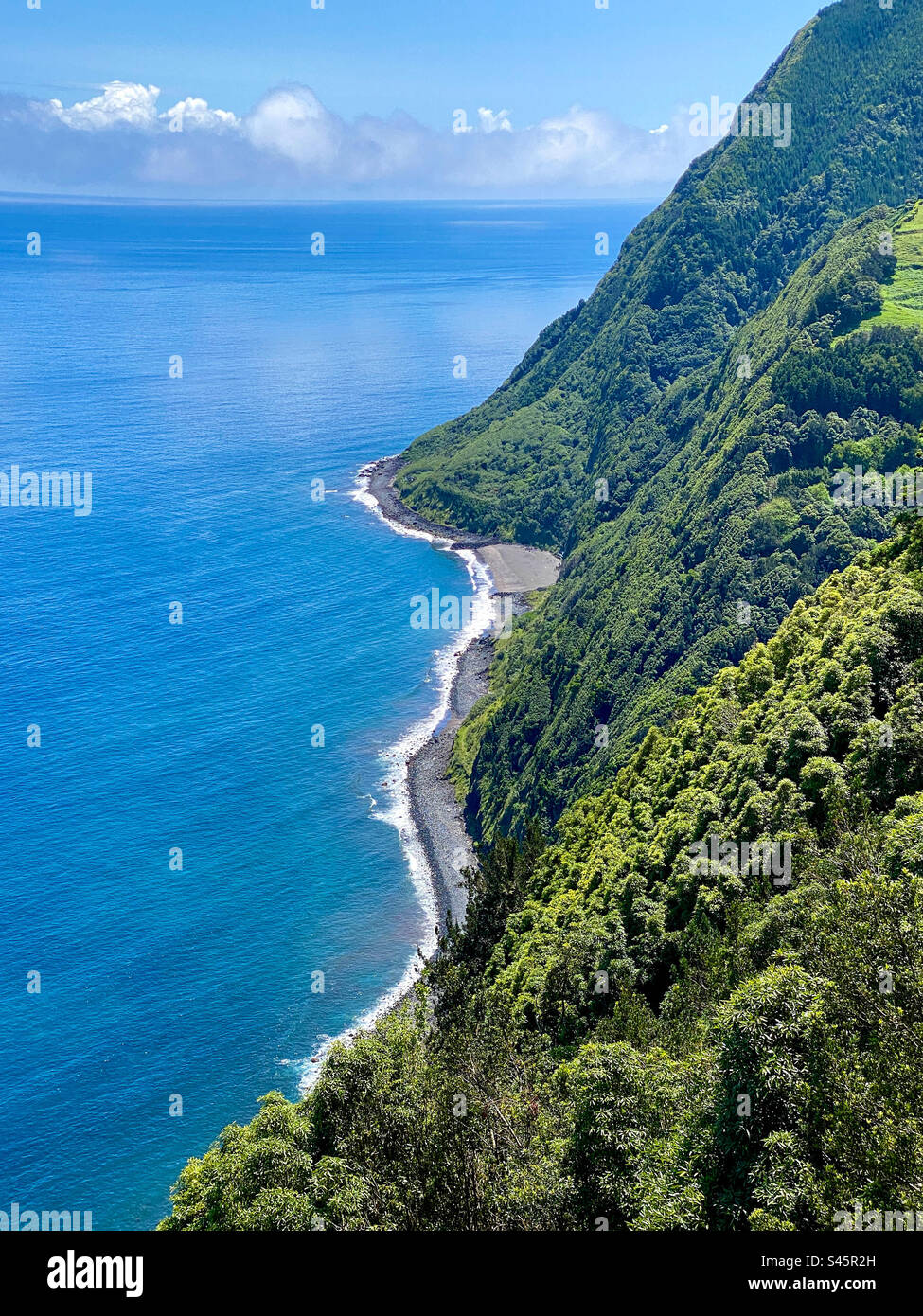 A rocky beach hugs the Atlantic Ocean shoreline at the bottom of towering plant-laden cliffs on Sao Miguel Island, Azores, Portugal - Smartphone Captured Stock Image