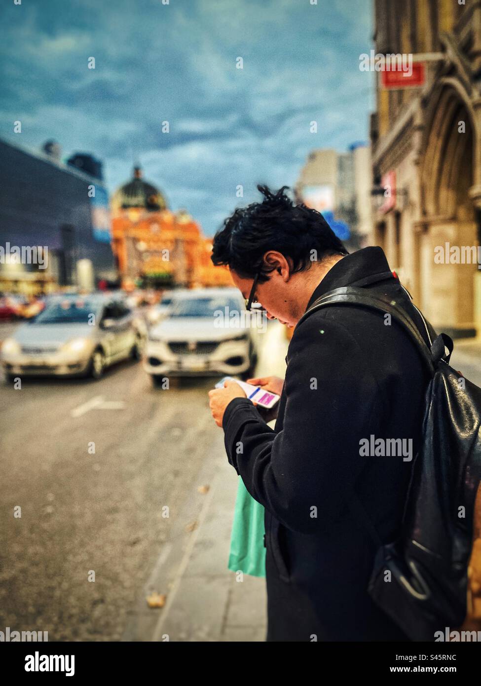 Young man using a mobile phone while standing on a sidewalk next to busy city road in Melbourne CBD, Victoria, Australia. Focus on foreground. Evening. - Smartphone Captured Stock Image