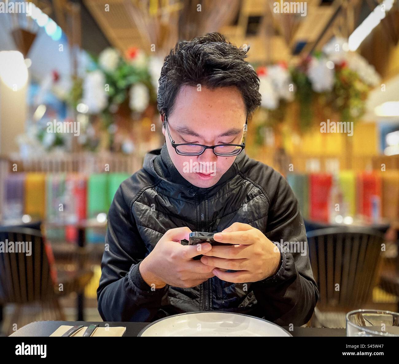Young Asian man in eyeglasses using mobile phone at table inside restaurant. Focus on foreground. - Smartphone Captured Stock Image