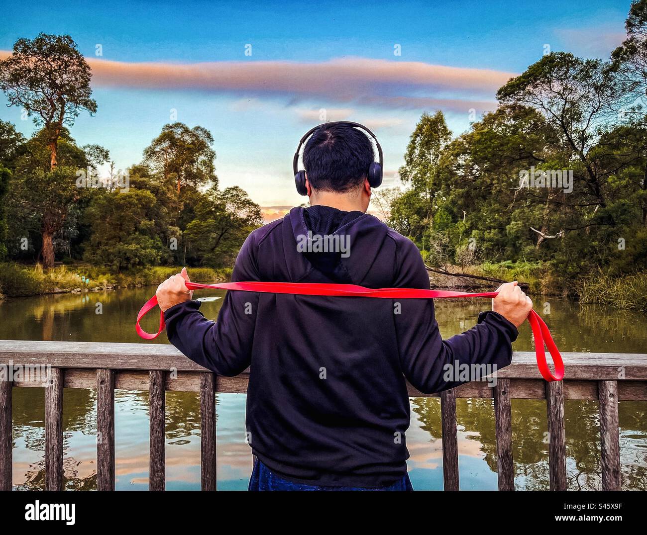 Rear view of young man exercising with resistance band in the park against sunset sky with pink cloud, trees and lake. Outdoor exercise. Tranquil scene. - Smartphone Captured Stock Image