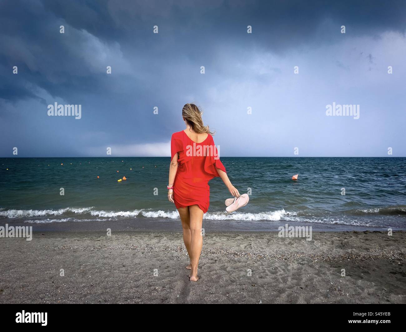 Rear view of woman in red dress walking down the beach towards the ocean on a cloudy day - Smartphone Captured Stock Image