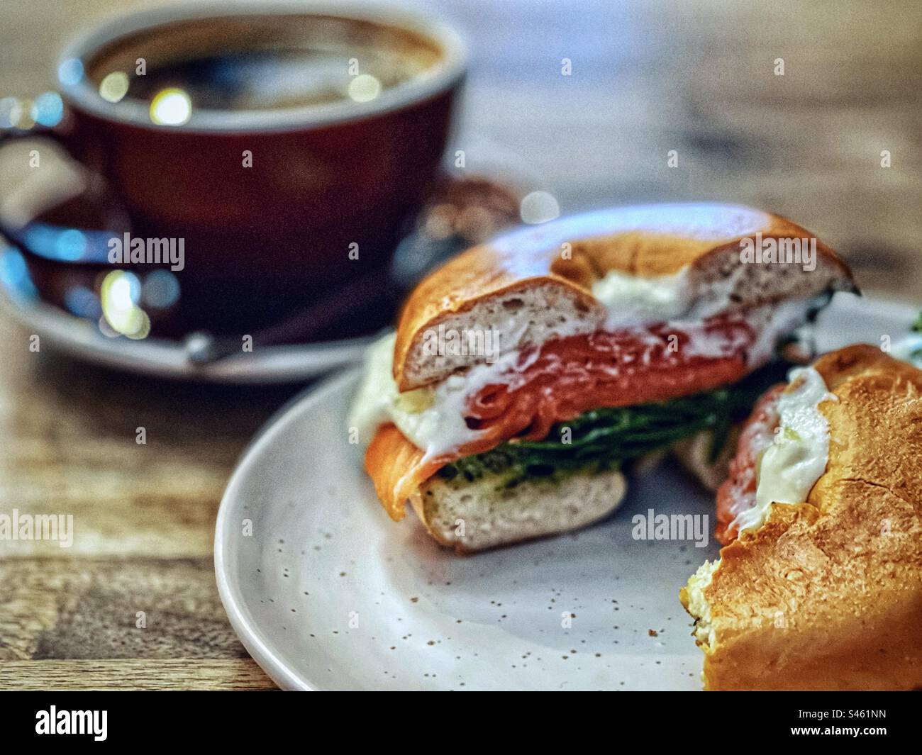 Close-up of salmon bagel with smoked salmon, spinach and tzatziki dip on plate and a cup of long black coffee on wooden table. - Smartphone Captured Stock Image