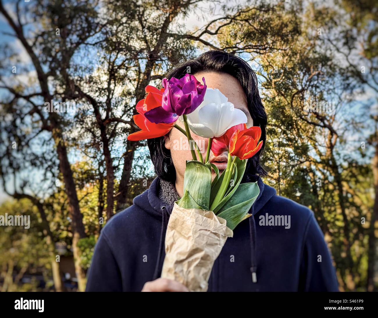 Portrait of young man holding a bouquet of four multicolored tulip flowers against trees and blue sky. Obscured face. Spring theme. Stock Photo