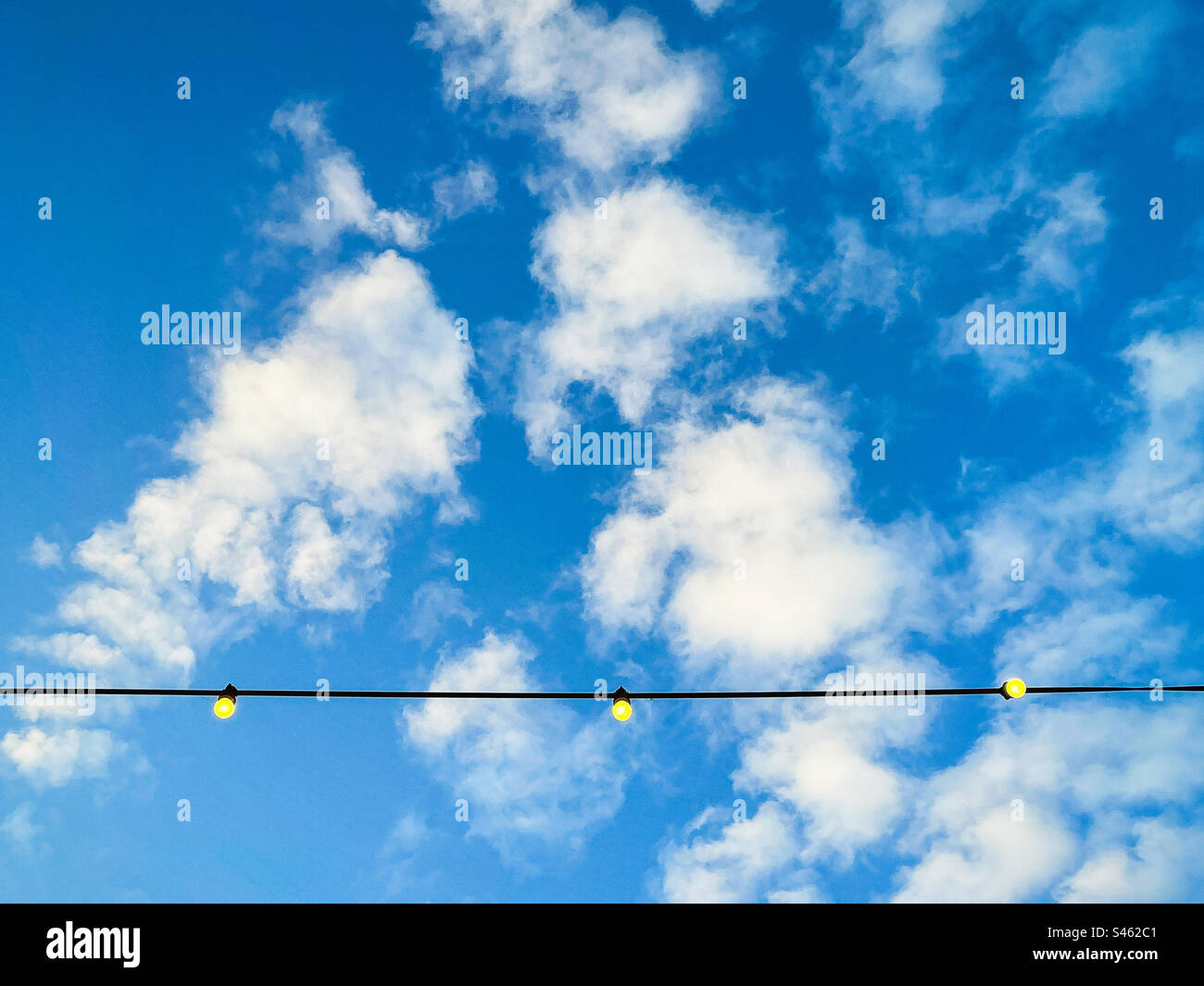 Low angle view of string of glowing light bulbs against blue sky with fluffy white clouds with copy space. - Smartphone Captured Stock Image