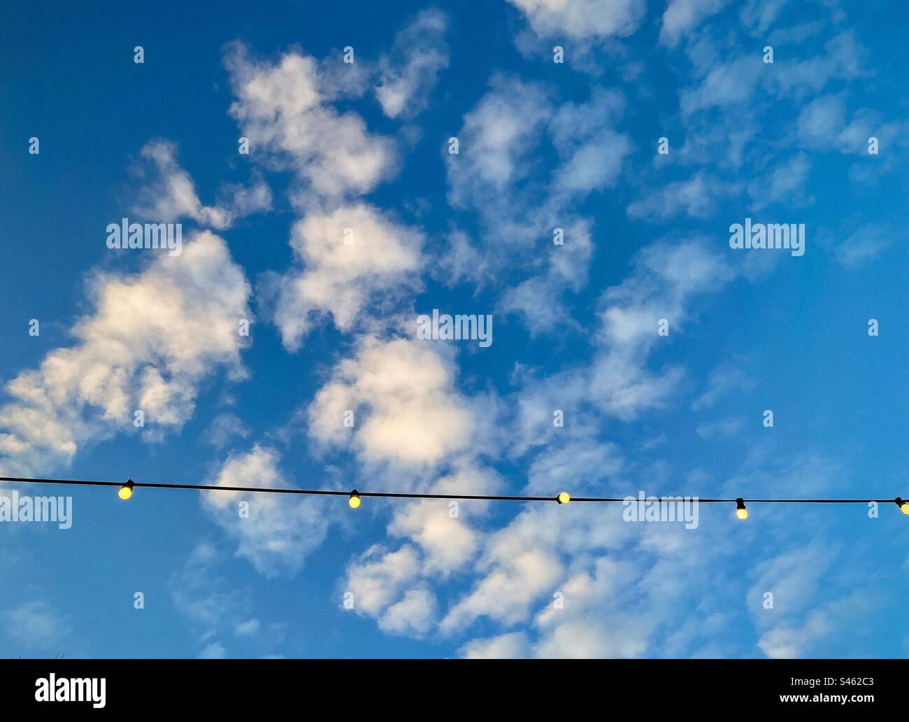 Low angle view of glowing light bulbs hanging from overhead line against blue sky with fluffy white clouds with copy space. String of light bulbs. - Smartphone Captured Stock Image
