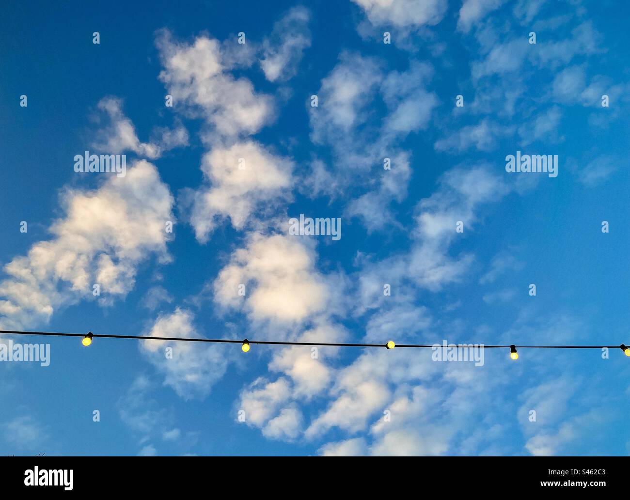 Low angle view of glowing light bulbs hanging from overhead line against blue sky with fluffy white clouds with copy space. String of light bulbs. Stock Photo