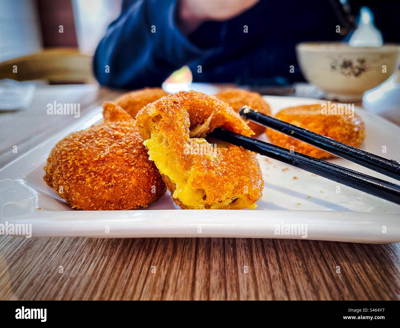 Close-up of fried pumpkin pastry balls with chopsticks on white plate against midsection of man at table. Focus on foreground. Chinese dessert. Yum cha dish. - Smartphone Captured Stock Image