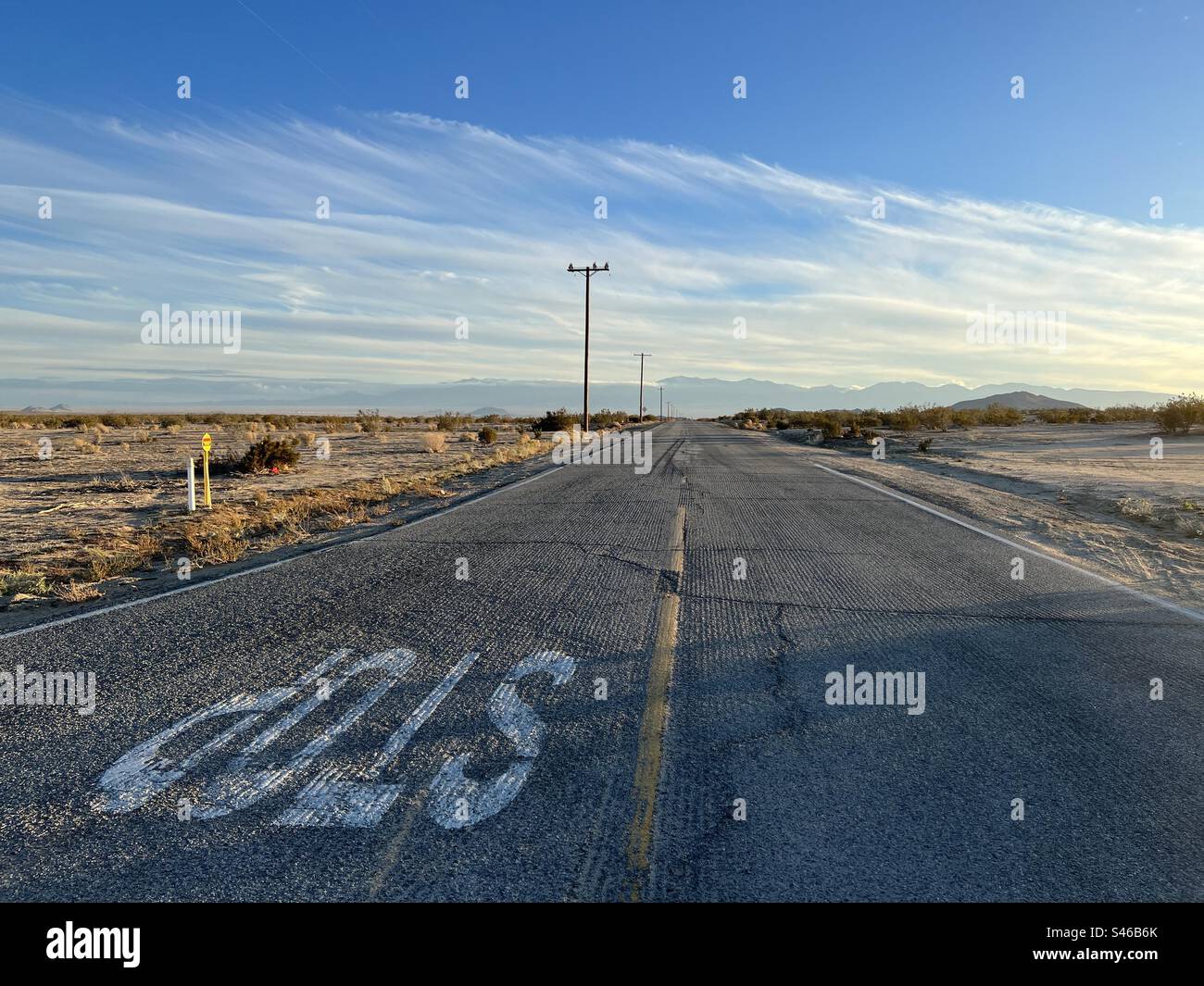 Stop sign on desert highway at traffic intersection in Southern California. Telephone poles with cables cross the road. Mountains visible in distance - Smartphone Captured Stock Image