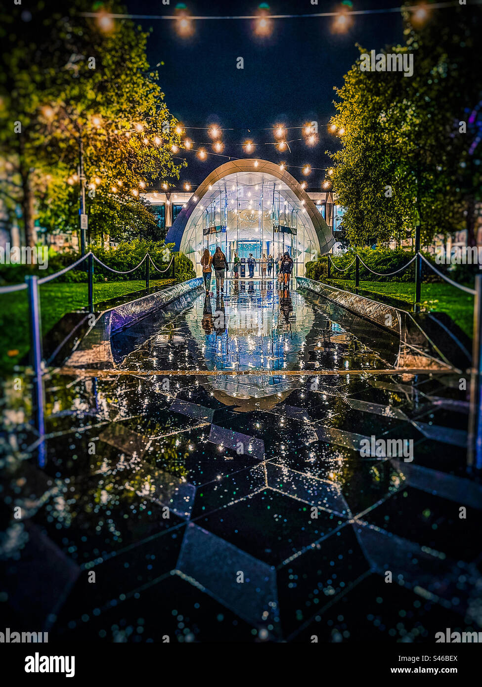 Groups of people entering and leaving the illuminated entrance of a shopping mall on a wet, rainy night. Reflections, illumination, night. - Smartphone Captured Stock Image