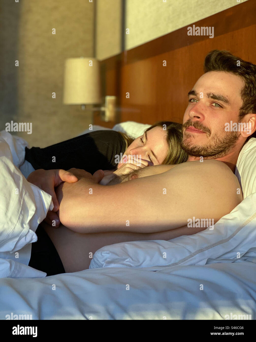 Young heterosexual couple snuggle in hotel room bed. Woman sleeps while man looks away from camera. - Smartphone Captured Stock Image