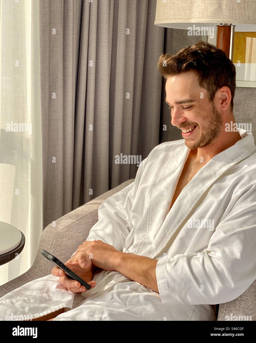 Handsome man dressed in white bathrobe smiles while he looks at his mobile phone. - Smartphone Captured Stock Image