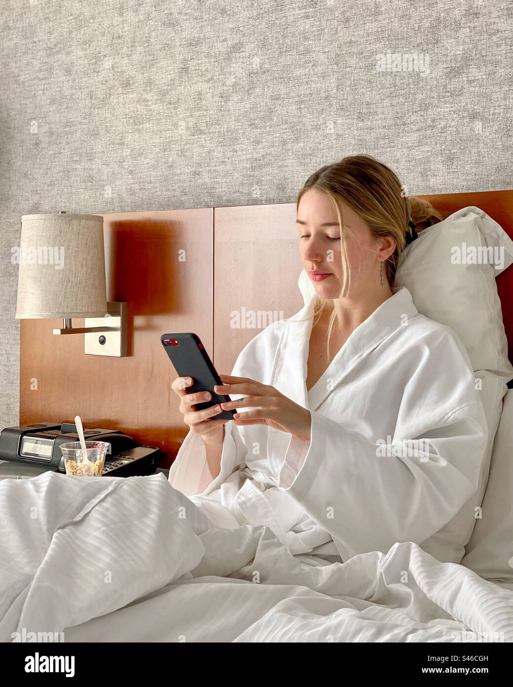 Young beautiful blonde woman dressed in bathrobe while sitting in hotel room bed using mobile phone. - Smartphone Captured Stock Image