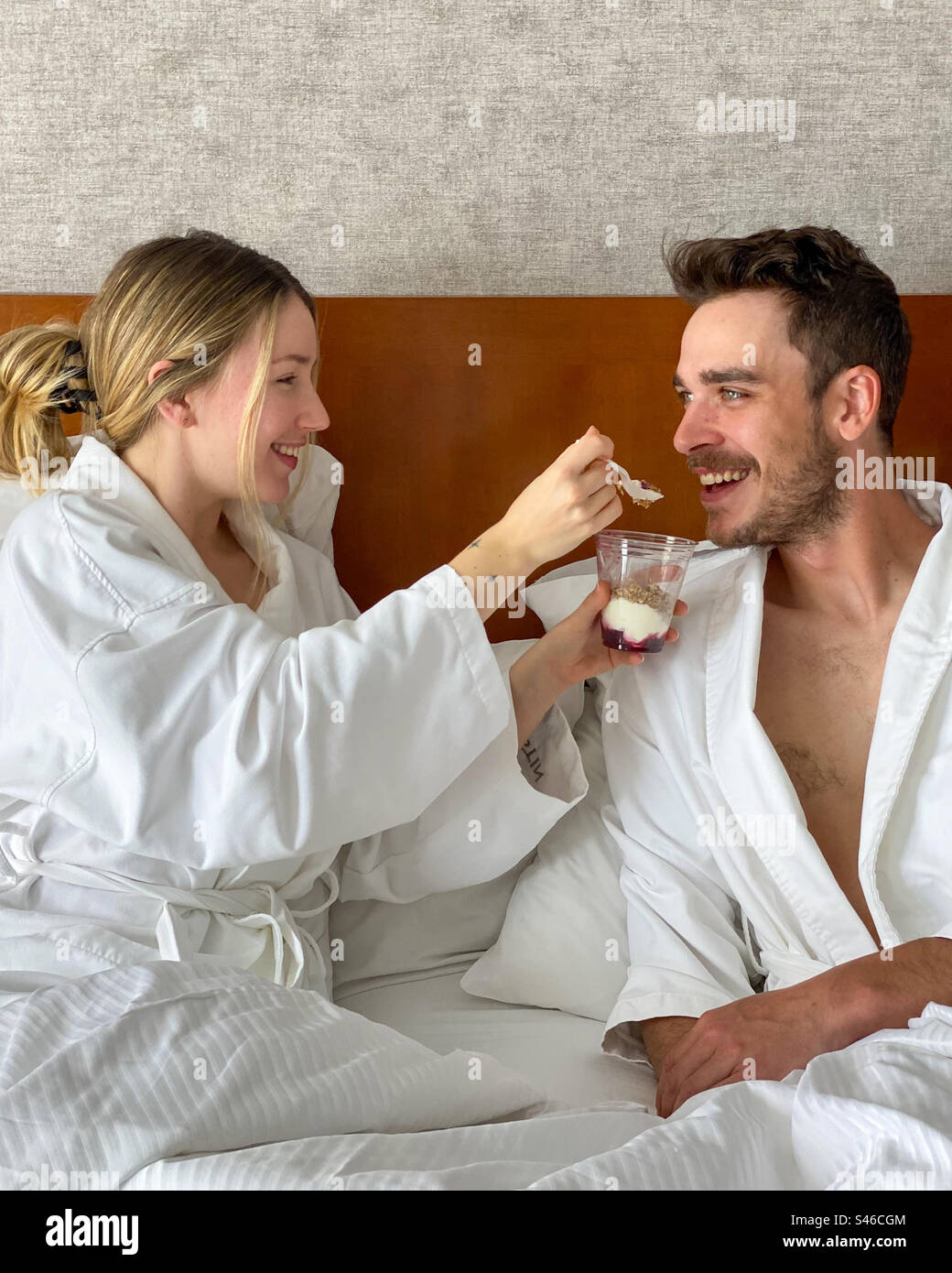 Young Caucasian couple dressed in white bathrobes I’m a hotel room bed. Woman shares her food with her partner. - Smartphone Captured Stock Image
