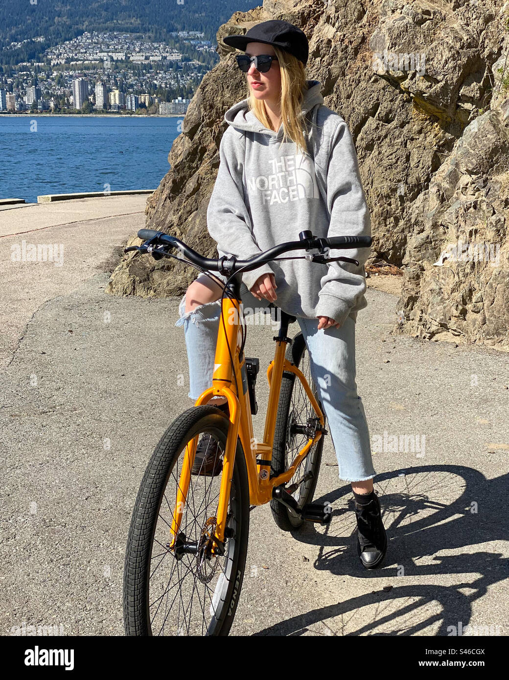 Young blonde hipster woman sits on a bicycle on a seaside path while looking away from the camera. - Smartphone Captured Stock Image