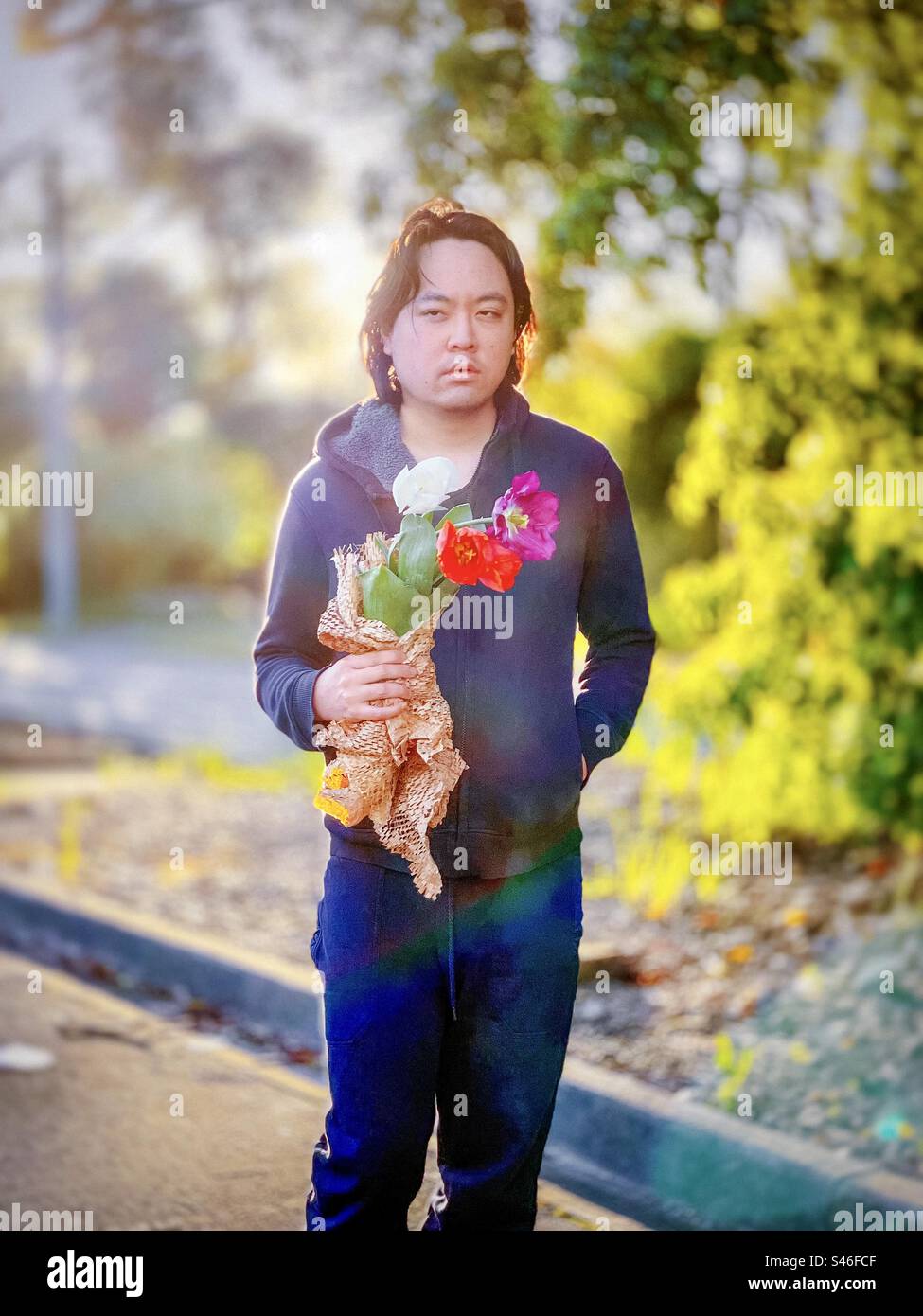 Three quarter length portrait of young Asian man holding white, red and purple tulips against setting sun and trees with rainbow lens flare. Focus on foreground. Spring theme. Golden hour. Dreamy. Stock Photo