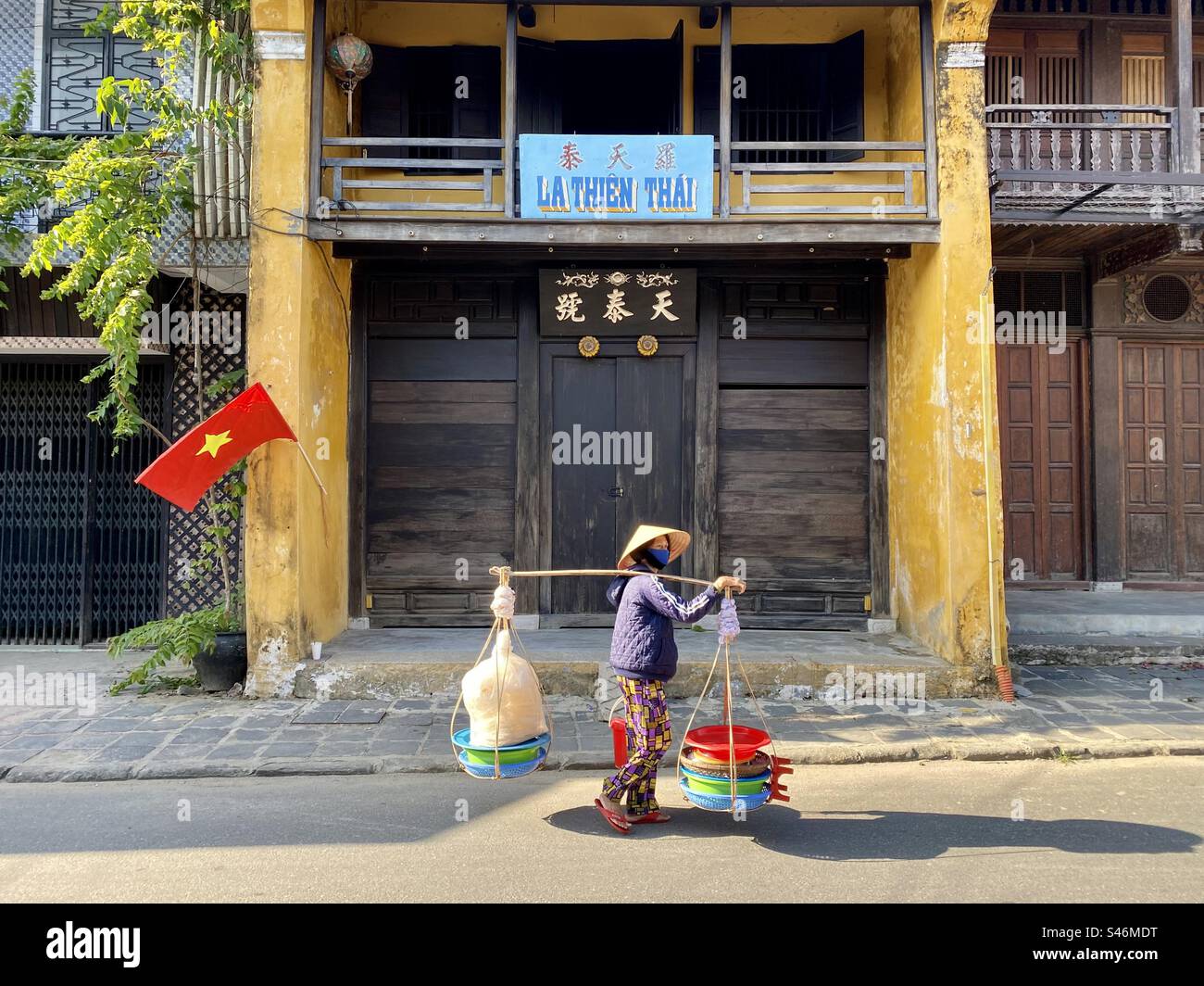 Vietnamese street vendor woman carrying goods in two baskets hanging at each end of a bamboo pole. Stock Photo