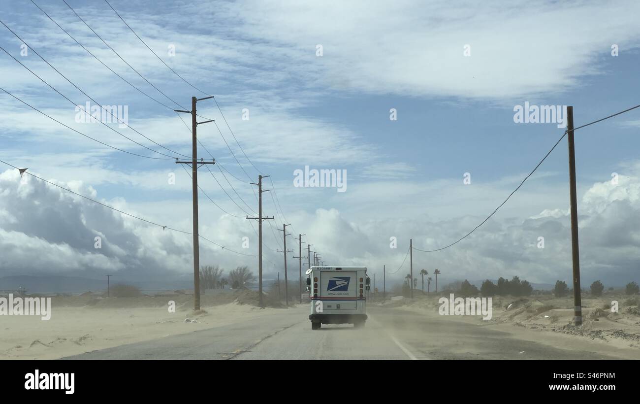 United States Postal Service van drives along road with dust storm on desert highway in southern California, with telegraph poles at side, fading into distance Stock Photo