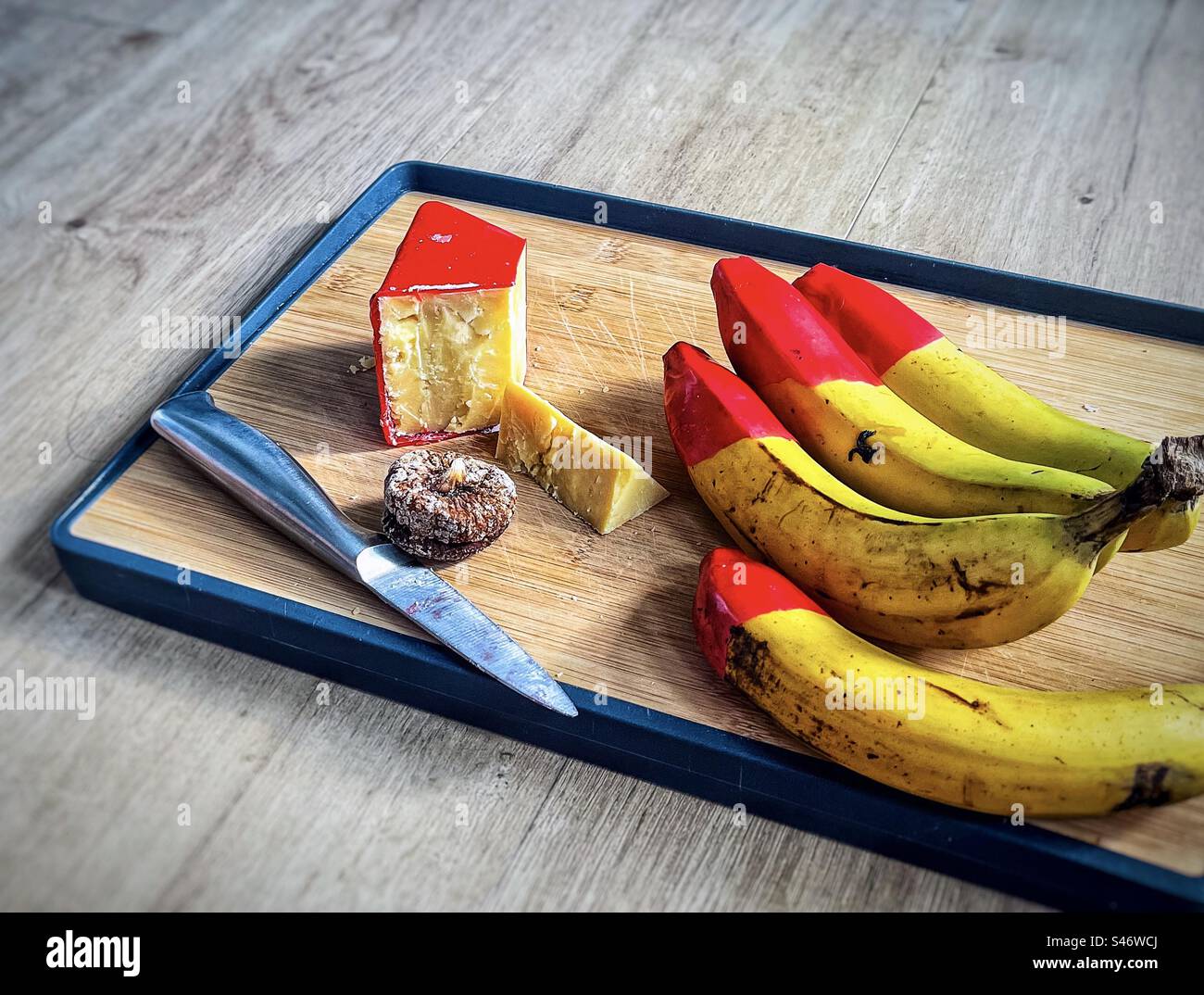 High angle view of aged cheddar cheese in red wax coating, red waxed tipped organic bananas, a dried fig and a knife on wooden cutting board on table with copy space. - Smartphone Captured Stock Image
