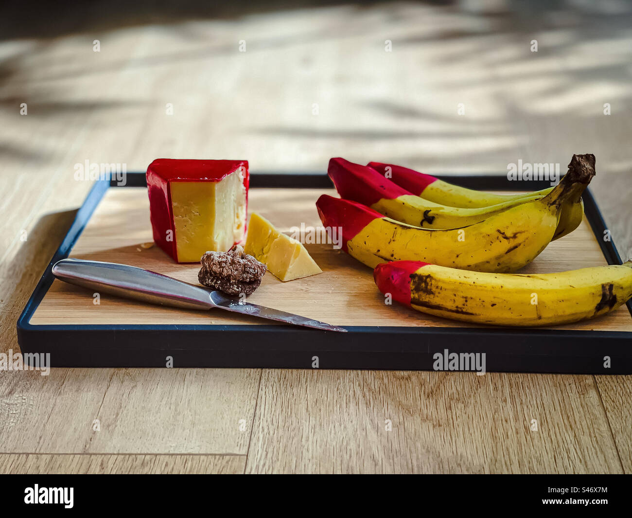 Aged cheddar cheese with red wax coating, a dried fig, organic, red wax tipped bananas and a knife on wooden cutting board on table with copy space. Sunlight and shadows. - Smartphone Captured Stock Image